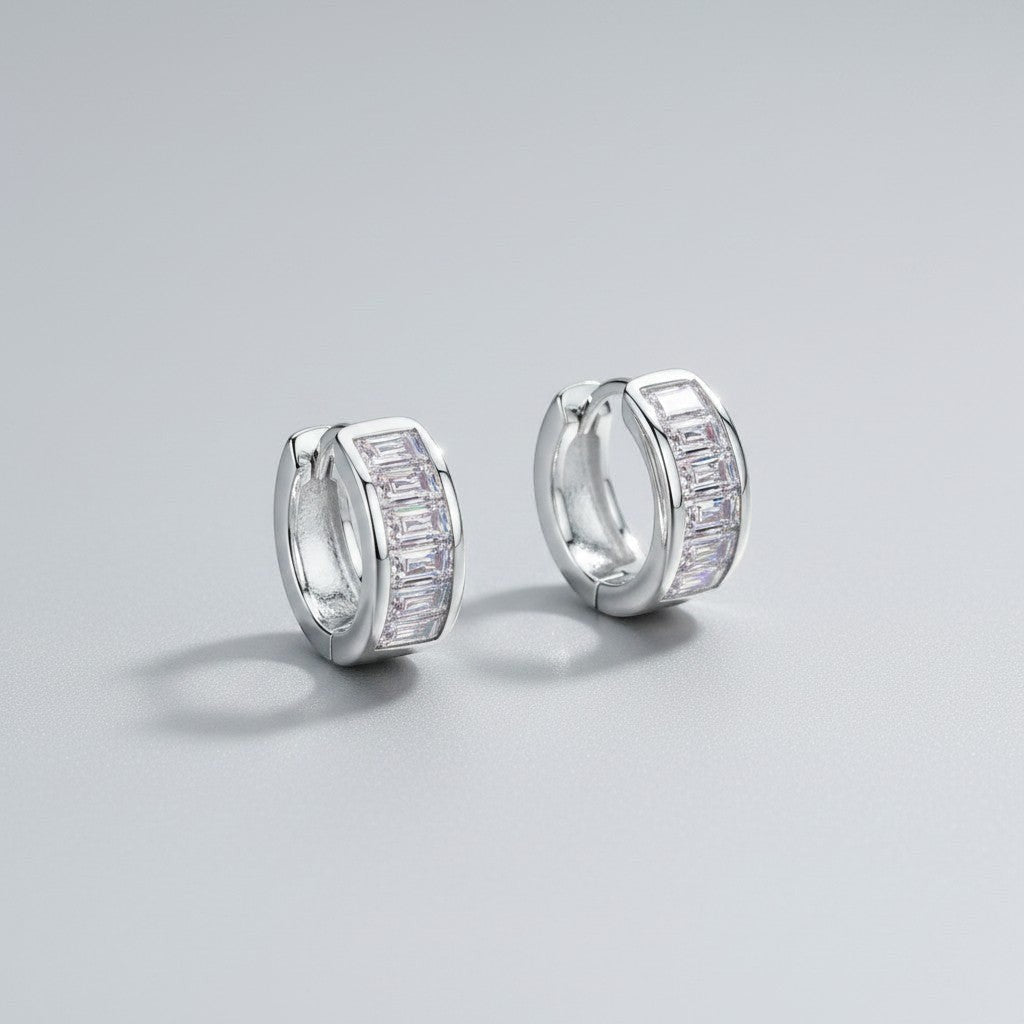 Schuinaanzicht van de twee rechtopstaande ModaMelhor 1.8 karaat baguette moissanite huggie oorbellen van gerhodineerd 925 sterling zilver met de focus op de glinsterende steentjes.