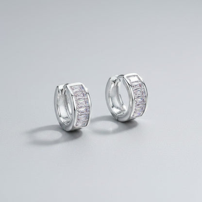 Schuinaanzicht van de twee rechtopstaande ModaMelhor 1.8 karaat baguette moissanite huggie oorbellen van gerhodineerd 925 sterling zilver met de focus op de glinsterende steentjes.