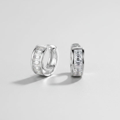 Vooraanzicht en zijaanzicht van de platliggende ModaMelhor 1.8 karaat baguette moissanite huggie oorbellen van gerhodineerd 925 sterling zilver met een strakke kanaalzetting.