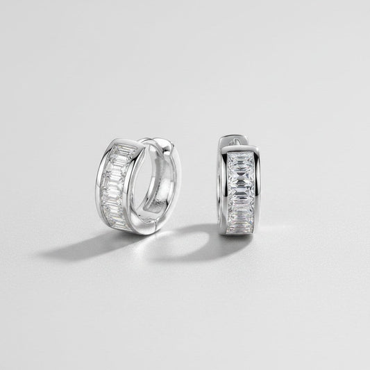 Vooraanzicht en zijaanzicht van de platliggende ModaMelhor 1.8 karaat baguette moissanite huggie oorbellen van gerhodineerd 925 sterling zilver met een strakke kanaalzetting.