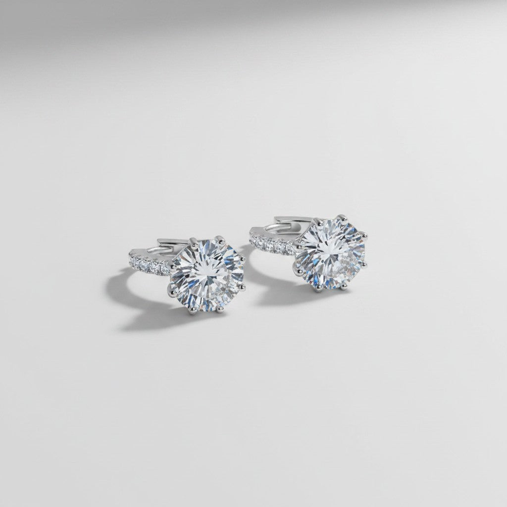 Schuinaanzicht van de twee platliggende ModaMelhor 4 karaat solitaire moissanite huggie oorbellen van gerhodineerd 925 sterling zilver met een opvallende ronde centrale steen en zirkonia accenten op de ring.