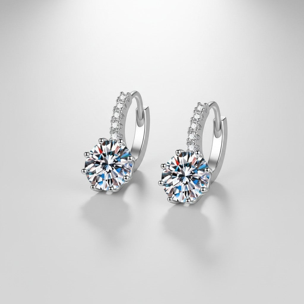 Vooraanzicht van de twee rechtopstaande ModaMelhor 4 karaat solitaire moissanite huggie oorbellen van gerhodineerd 925 sterling zilver met de focus op de grote centrale stenen en zirkonia accenten.