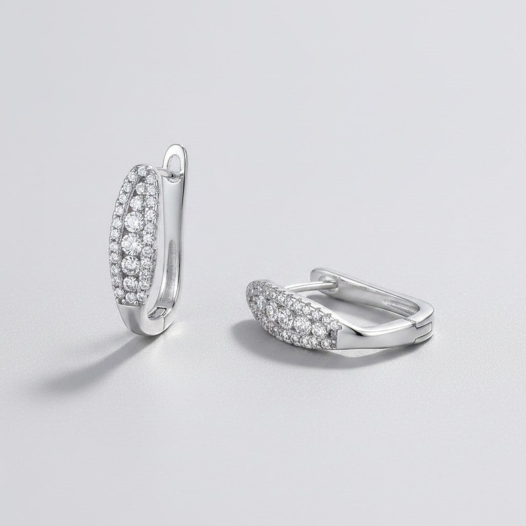 Vooraanzicht en zijaanzicht van de platliggende ModaMelhor ovalen moissanite huggie oorbellen van gerhodineerd 925 sterling zilver bezet met meerdere rijen kleine moissanite stenen.