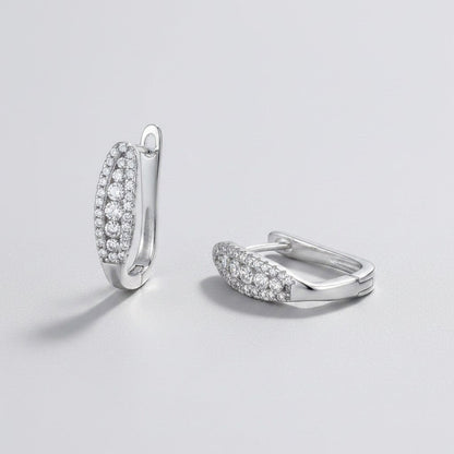 Vooraanzicht en zijaanzicht van de platliggende ModaMelhor ovalen moissanite huggie oorbellen van gerhodineerd 925 sterling zilver bezet met meerdere rijen kleine moissanite stenen.