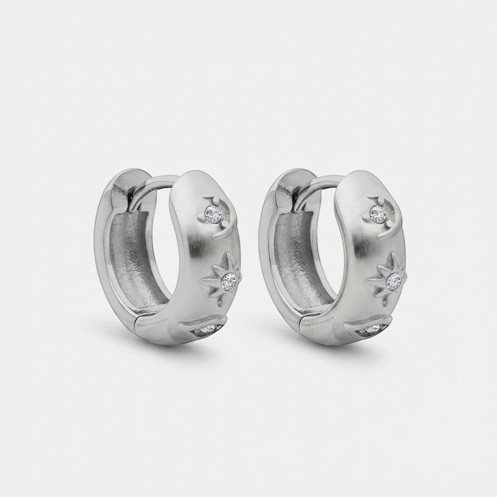 Schuinaanzicht van de twee rechtopstaande ModaMelhor titanium huggie oorbellen van zilverkleurig titanium, waarop het motief met zirkonia steentjes aan de zijkant duidelijk zichtbaar is.