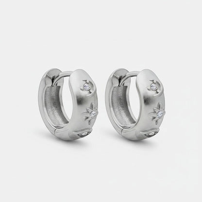 Schuinaanzicht van de twee rechtopstaande ModaMelhor titanium huggie oorbellen van zilverkleurig titanium, waarop het motief met zirkonia steentjes aan de zijkant duidelijk zichtbaar is.