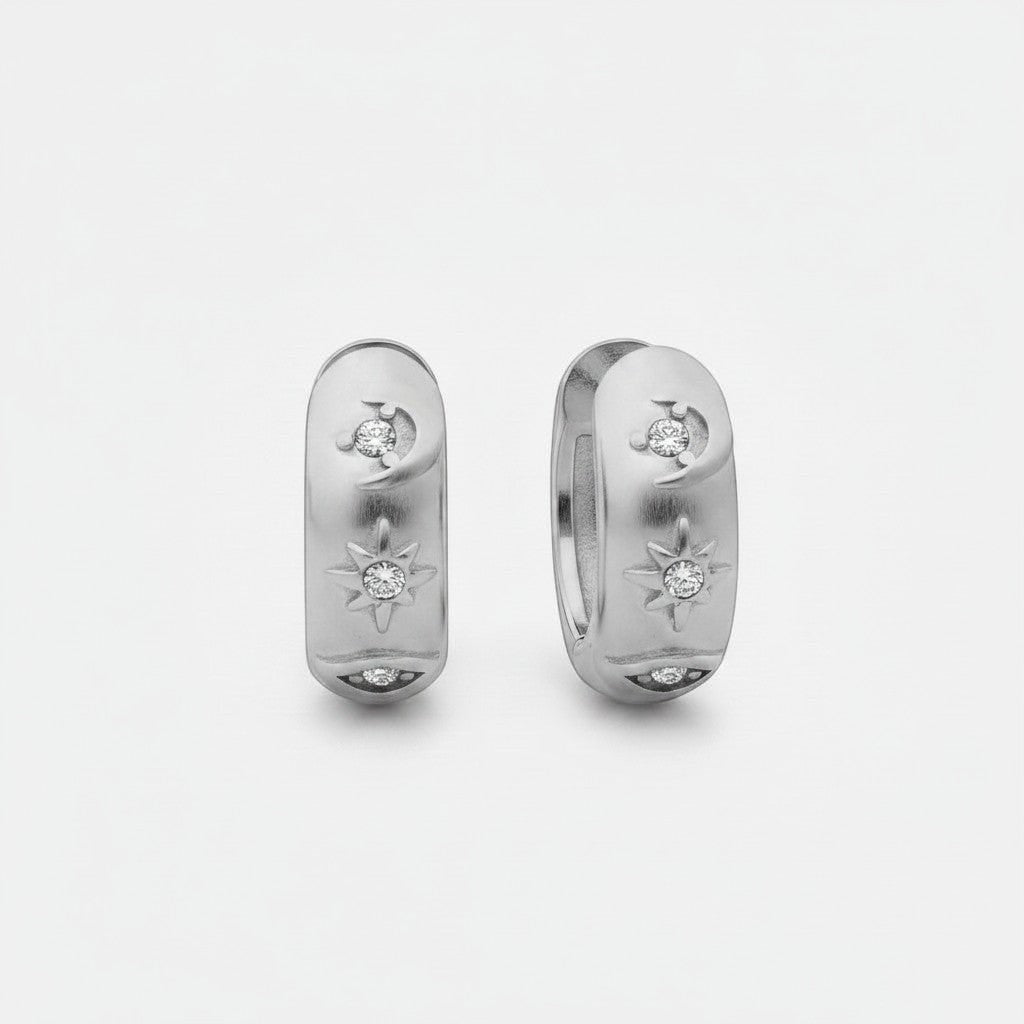 Vooraanzicht van de twee rechtopstaande ModaMelhor titanium huggie oorbellen van zilverkleurig titanium, voorzien van een motief met een maan, ster en oog ingelegd met zirkonia steentjes.