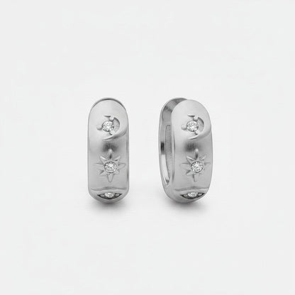 Vooraanzicht van de twee rechtopstaande ModaMelhor titanium huggie oorbellen van zilverkleurig titanium, voorzien van een motief met een maan, ster en oog ingelegd met zirkonia steentjes.