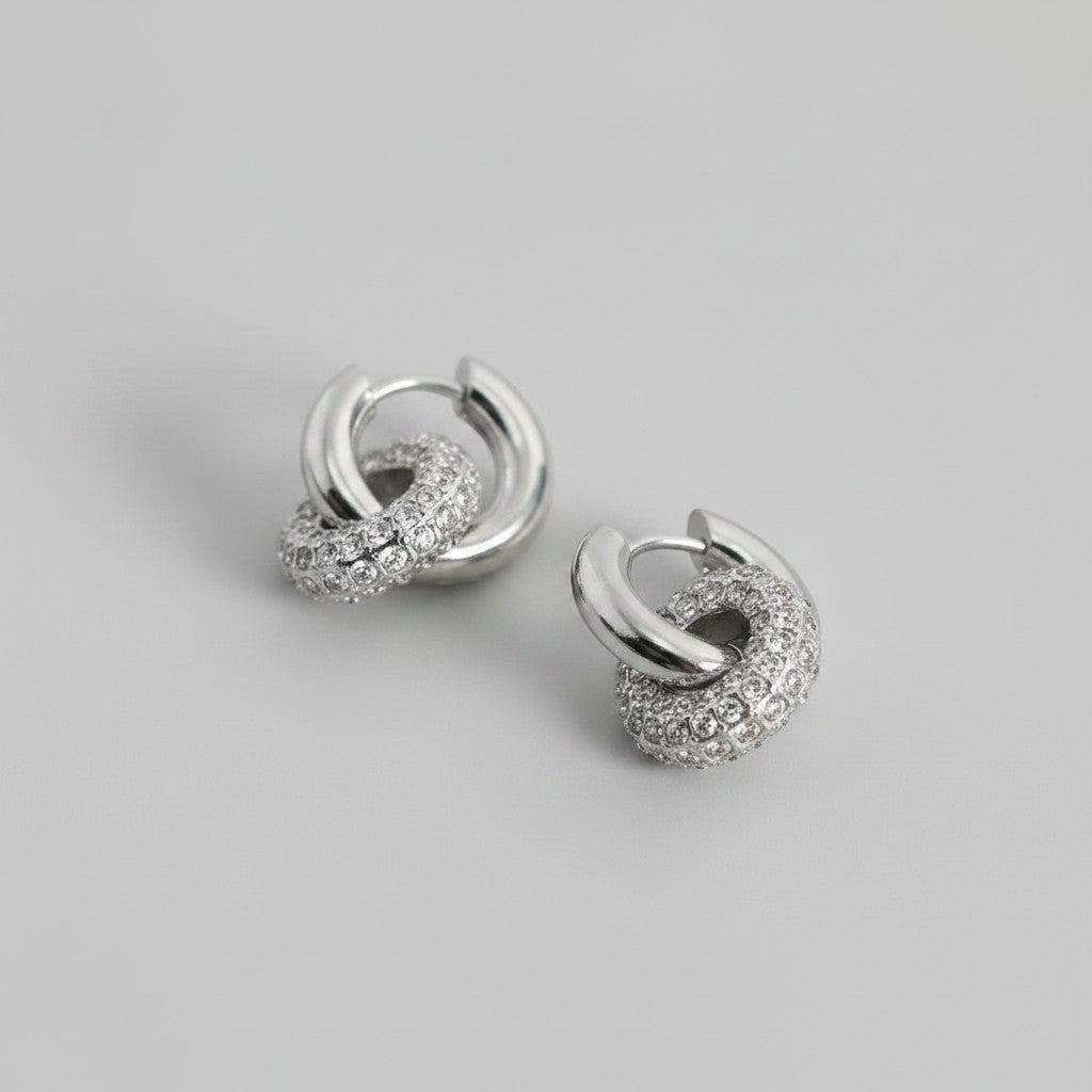 Schuinaanzicht van de twee platliggende ModaMelhor zirconia titanium double hoop oorbellen van zilverkleurig titanium, waarbij een gladde ring is verbonden met een ring vol zirkonia steentjes.