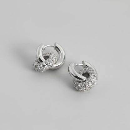 Schuinaanzicht van de twee platliggende ModaMelhor zirconia titanium double hoop oorbellen van zilverkleurig titanium, waarbij een gladde ring is verbonden met een ring vol zirkonia steentjes.